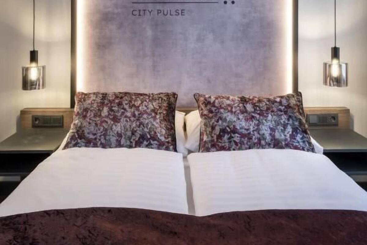 A bed with floral pillows, a large wall with 'andre CITY PULSE' text, and pendant lights.