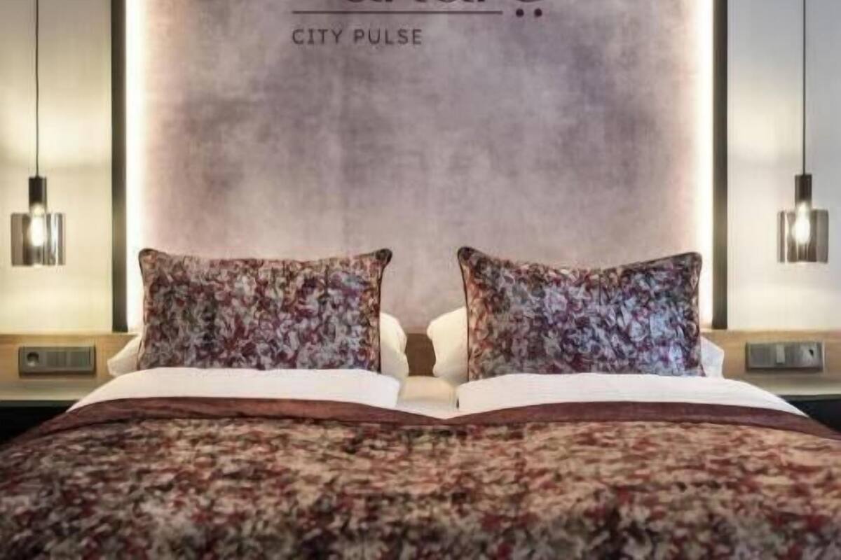 A hotel room with a large bed, two matching pillows, a wall-mounted light, and a sign reading 'andre CITY PULSE'.