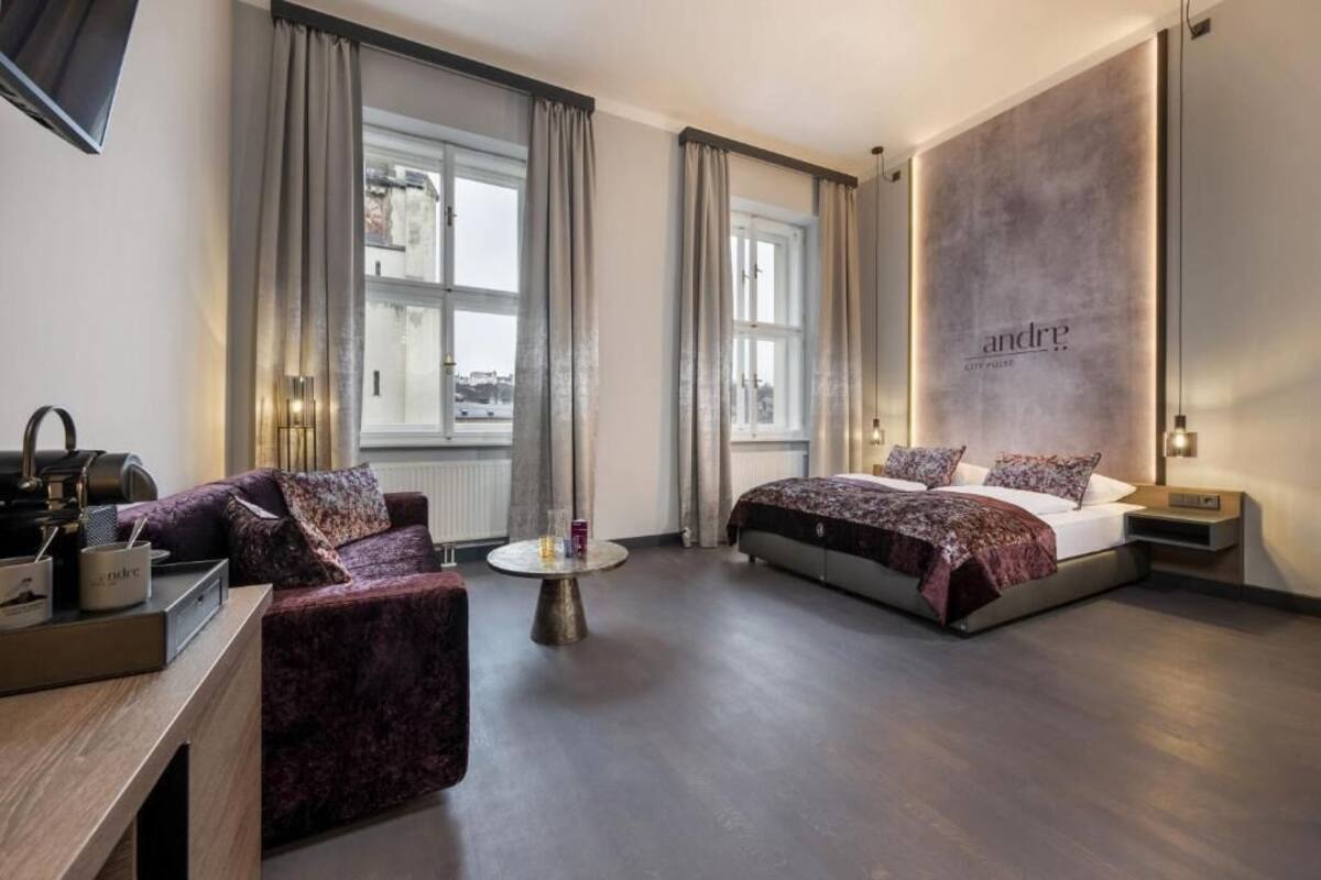 A modern hotel room with a bed, a sofa, a small round table, and a large window.