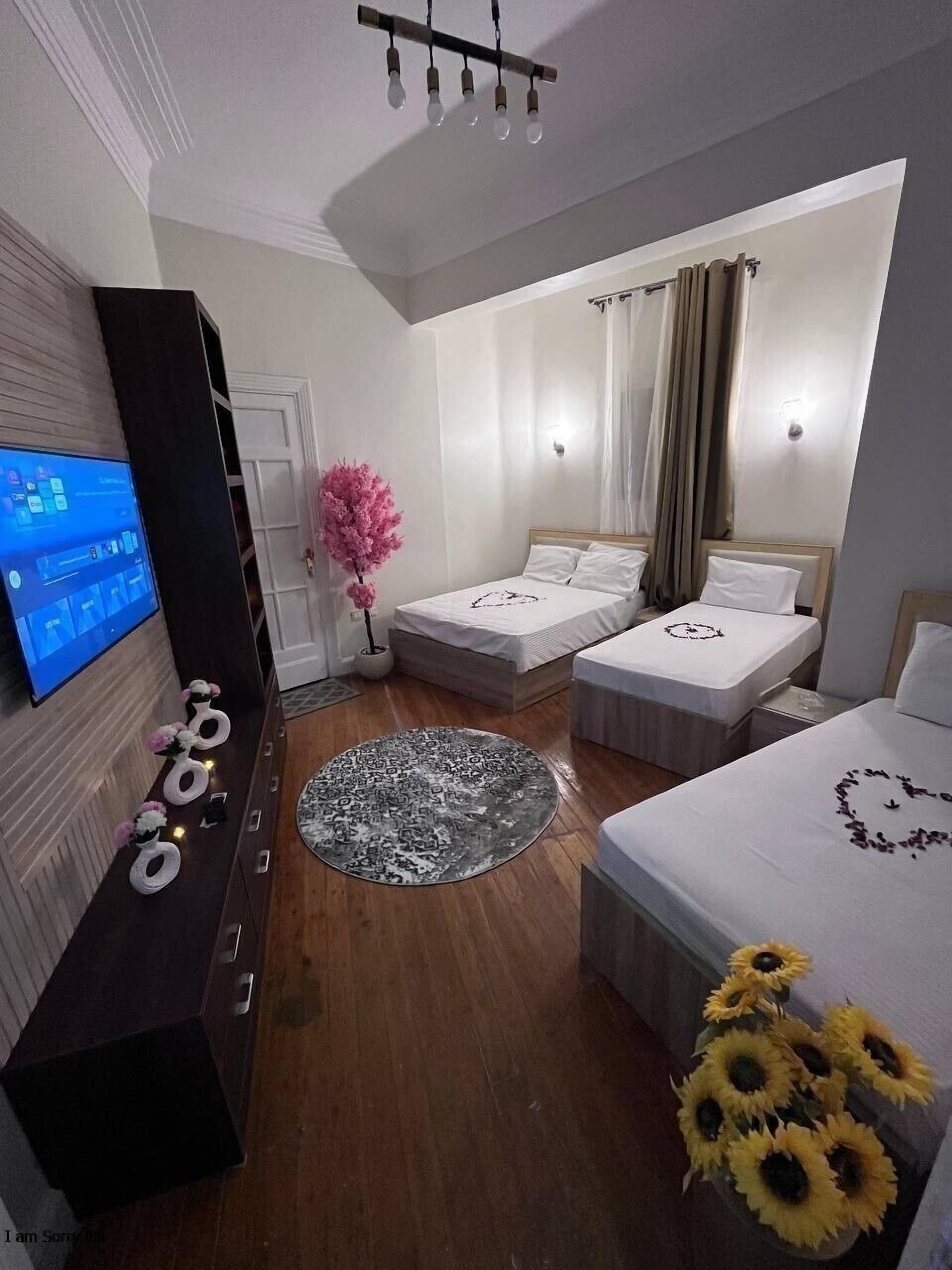 Una habitación de hotel moderna con dos camas individuales, un televisor montado en un mueble de madera oscura, un tapete redondo estampado y un jarrón con girasoles.