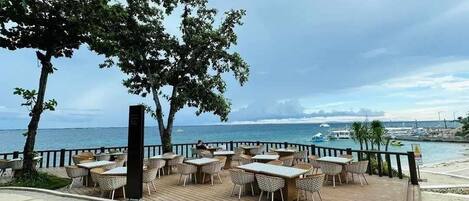 A seaside restaurant with outdoor seating, a wooden deck, and a view of the ocean and boats.
