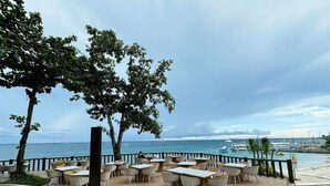 A seaside restaurant with outdoor seating, a wooden deck, and a view of the ocean and boats.