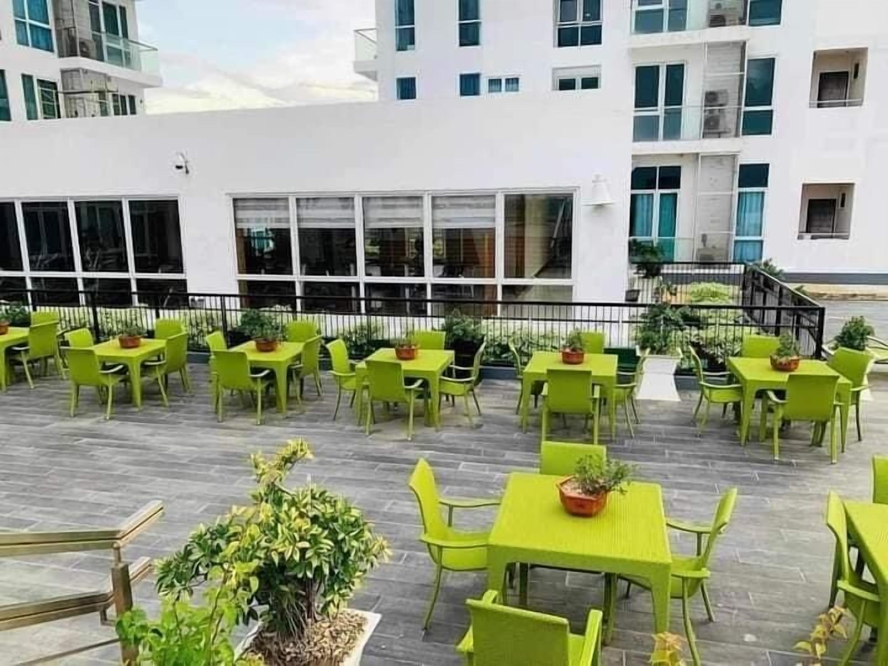 An outdoor seating area with green tables and chairs, surrounded by potted plants and a white building in the background.