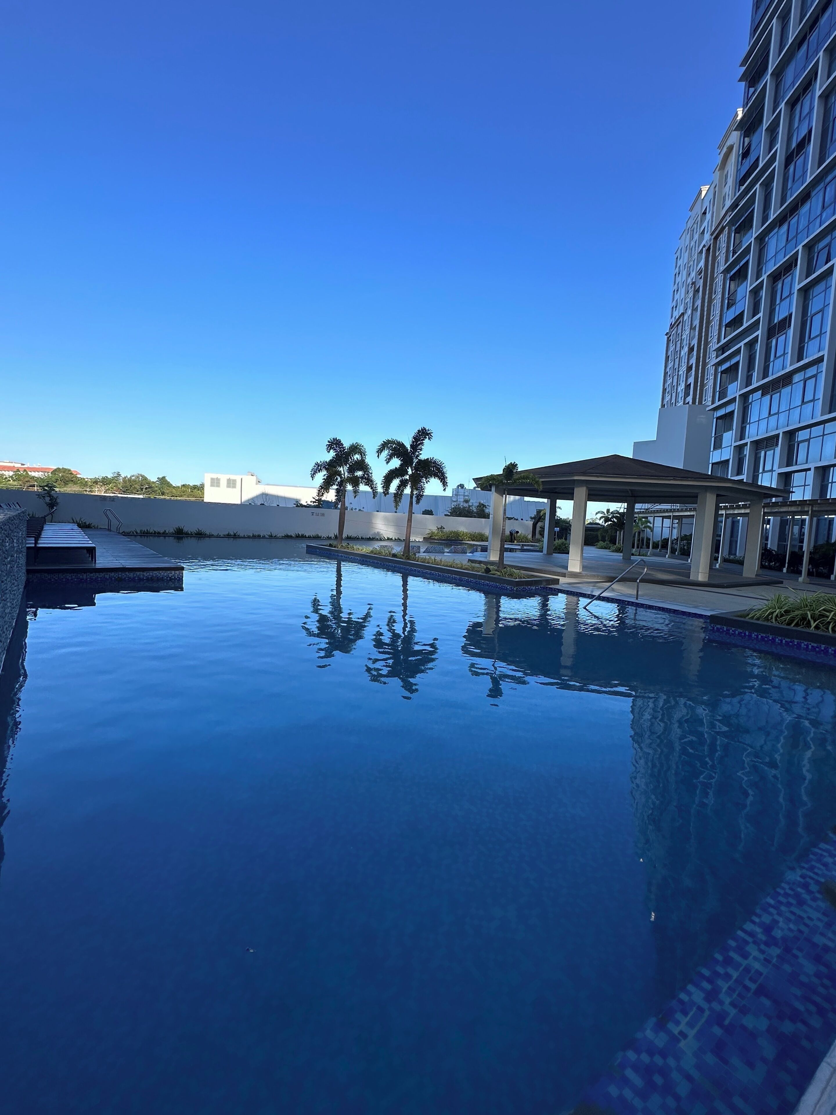 Un cielo azul despejado, una piscina con reflejos, un edificio moderno y palmeras.