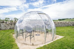 A transparent geodesic dome structure with a circular table and chairs inside, surrounded by a stone wall and grassy area. - The Duke - North East Escapes (Bamburgh)