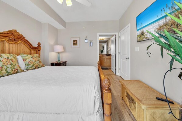 A bedroom with a large bed, a wooden headboard, a bedside table with a lamp, a small cabinet, and a wall-mounted TV. - Grandview East 1201 (Panama City Beach)
