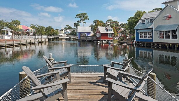 A wooden dock with chairs, a calm water body, and buildings with a rustic architectural style. - Augusta Village Sandestin by Gibson BR  (Miramar Beach)