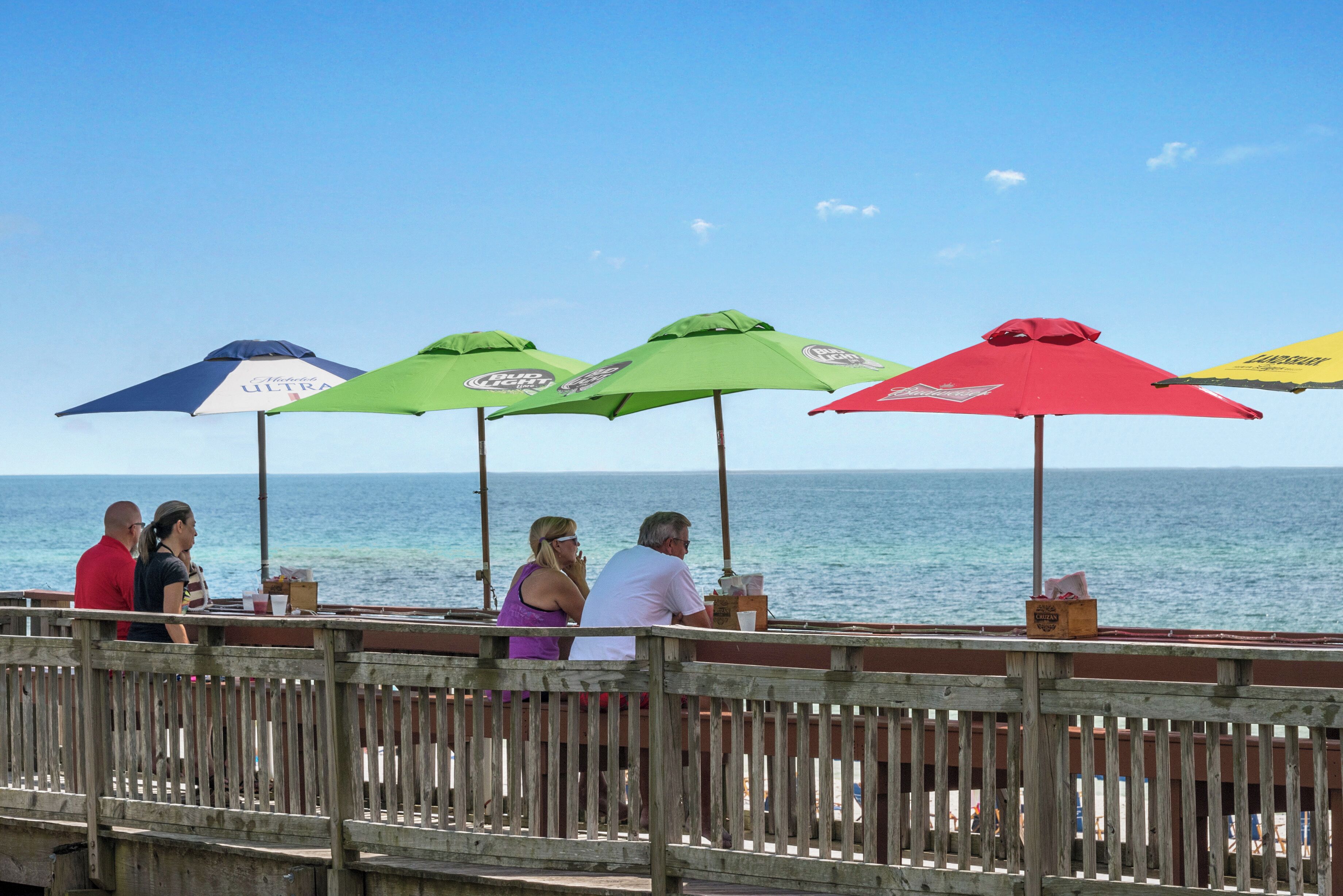 A wooden pier with people seated under colorful umbrellas, overlooking the sea.