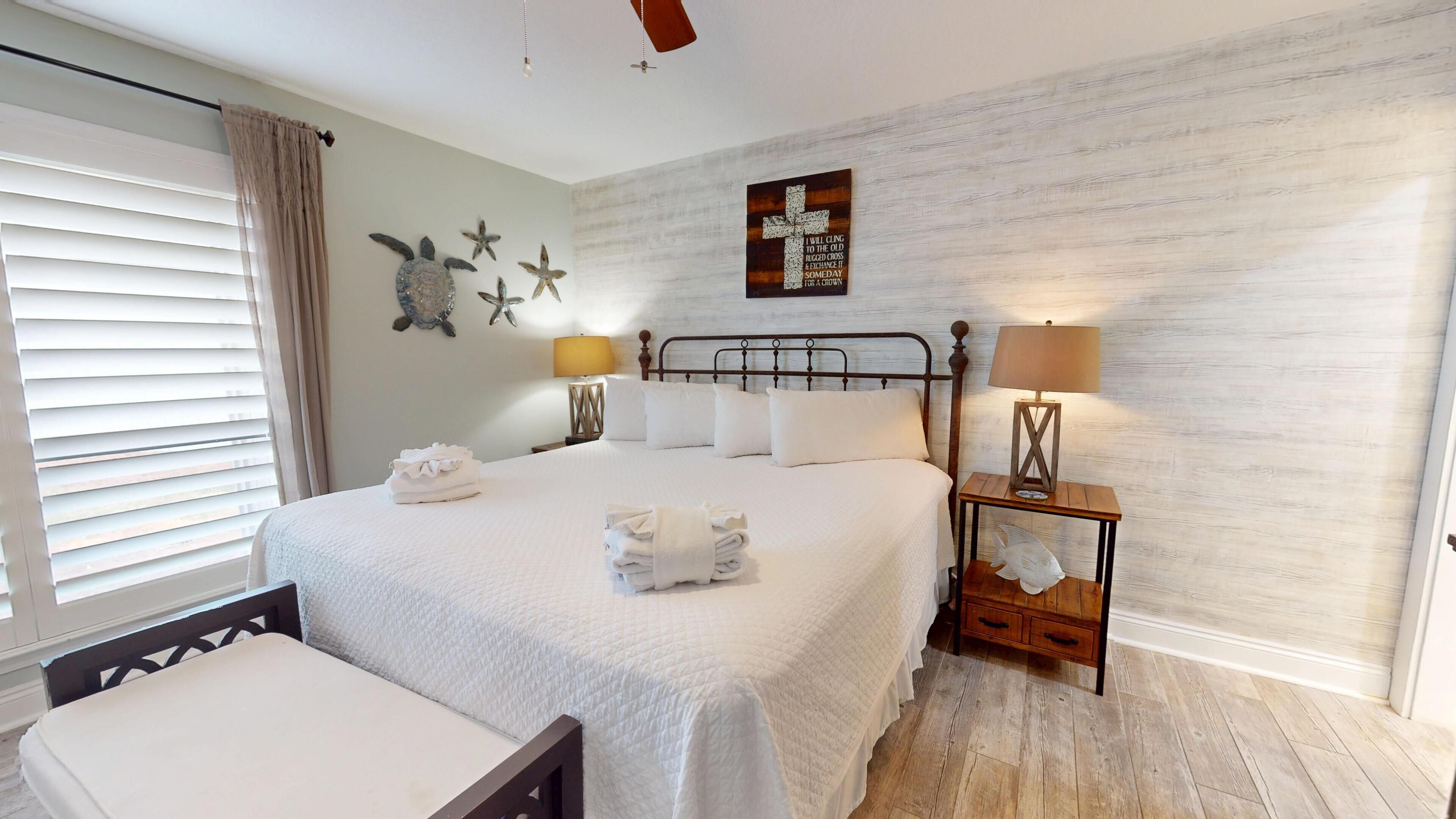 Cama bem arrumada com lençóis brancos, mesa de cabeceira com abajur e arte decorativa na parede.