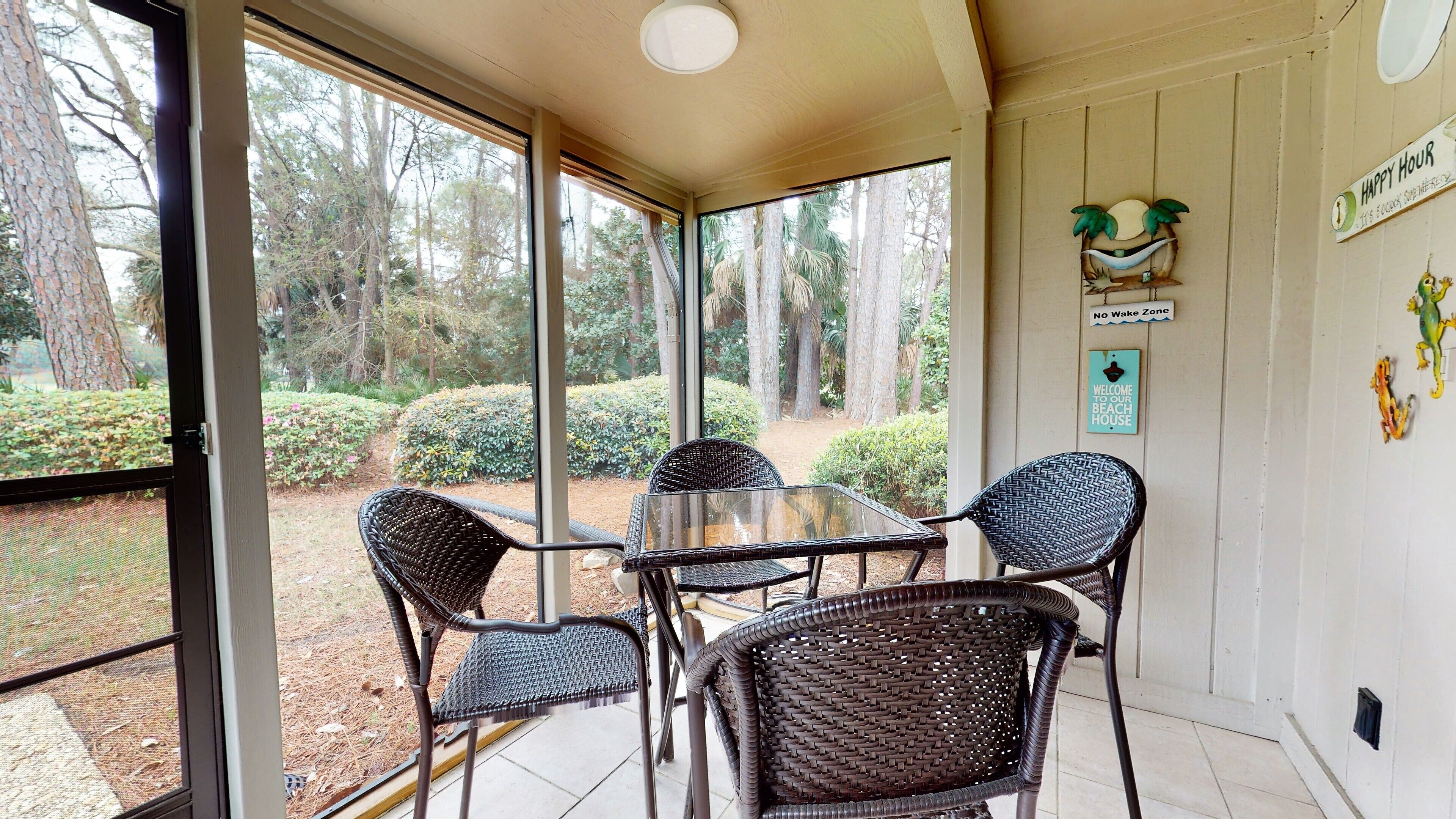 A covered patio with wicker chairs and a glass table, overlooking a landscaped garden with trees and shrubs.