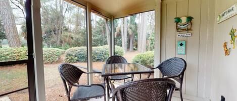 A covered patio with wicker chairs and a glass table, overlooking a landscaped garden with trees and shrubs.