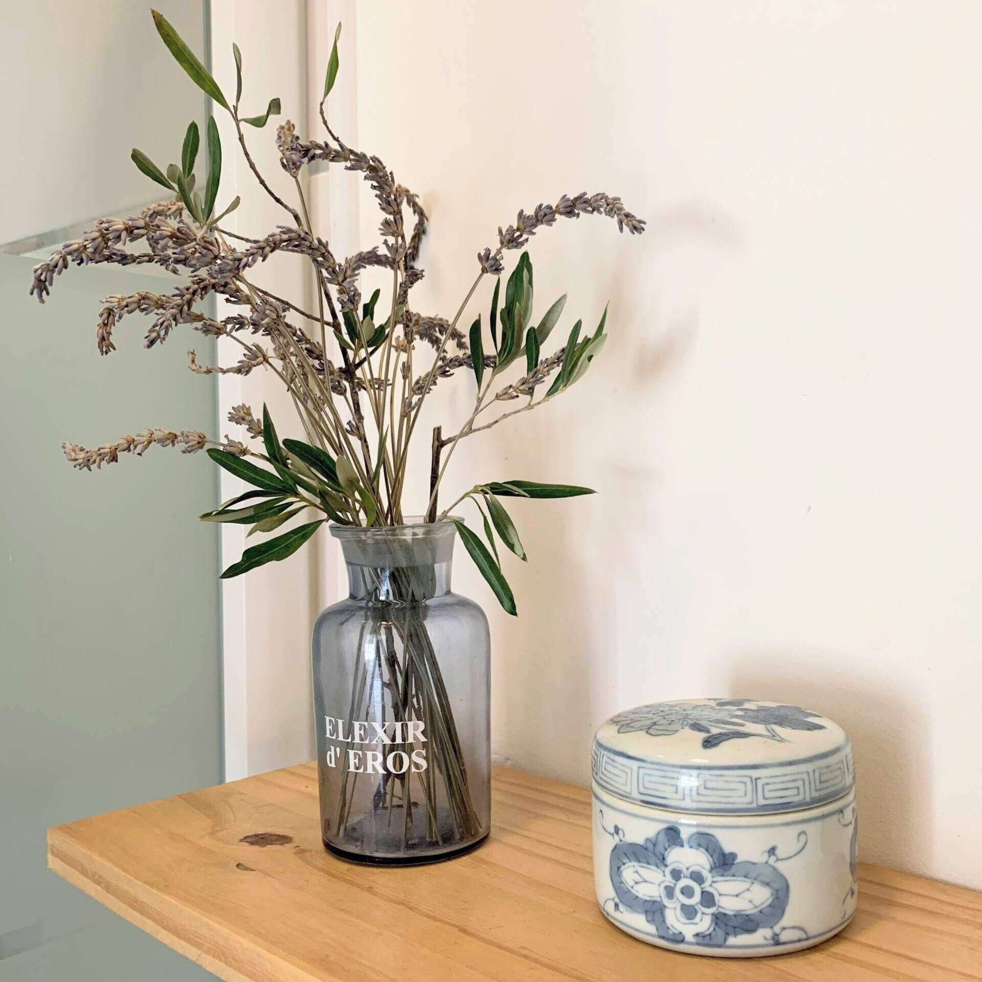 A clear glass vase with dried flowers and green leaves on a wooden shelf next to a ceramic container with blue floral designs.