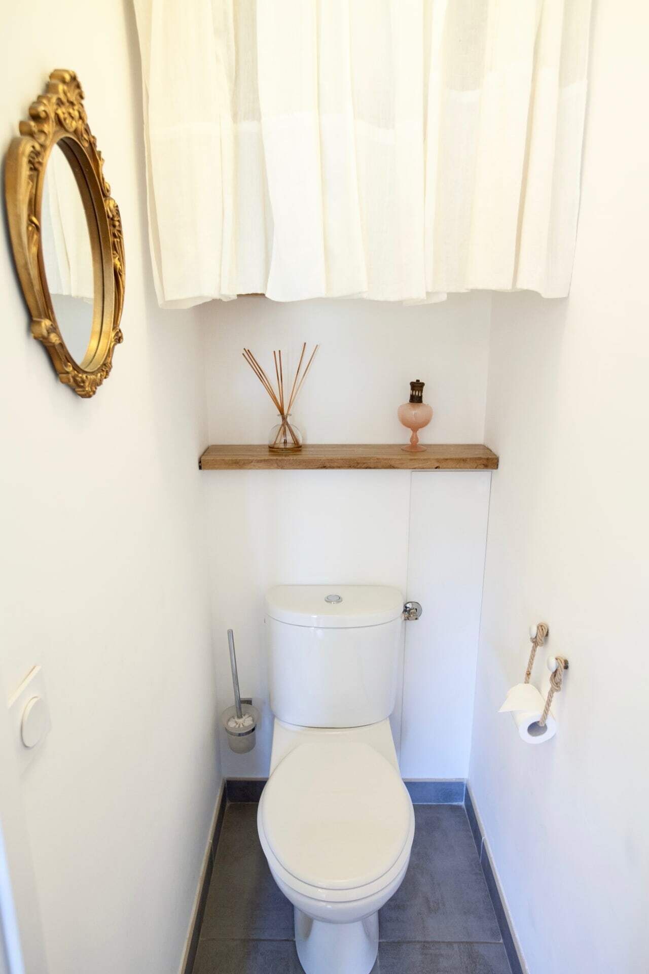 Ein kleines, sauberes Badezimmer mit Toilette, einem spiegel mit goldener Rahmen, einem Regal mit Dekoration und einem Toilettenpapierhalter.