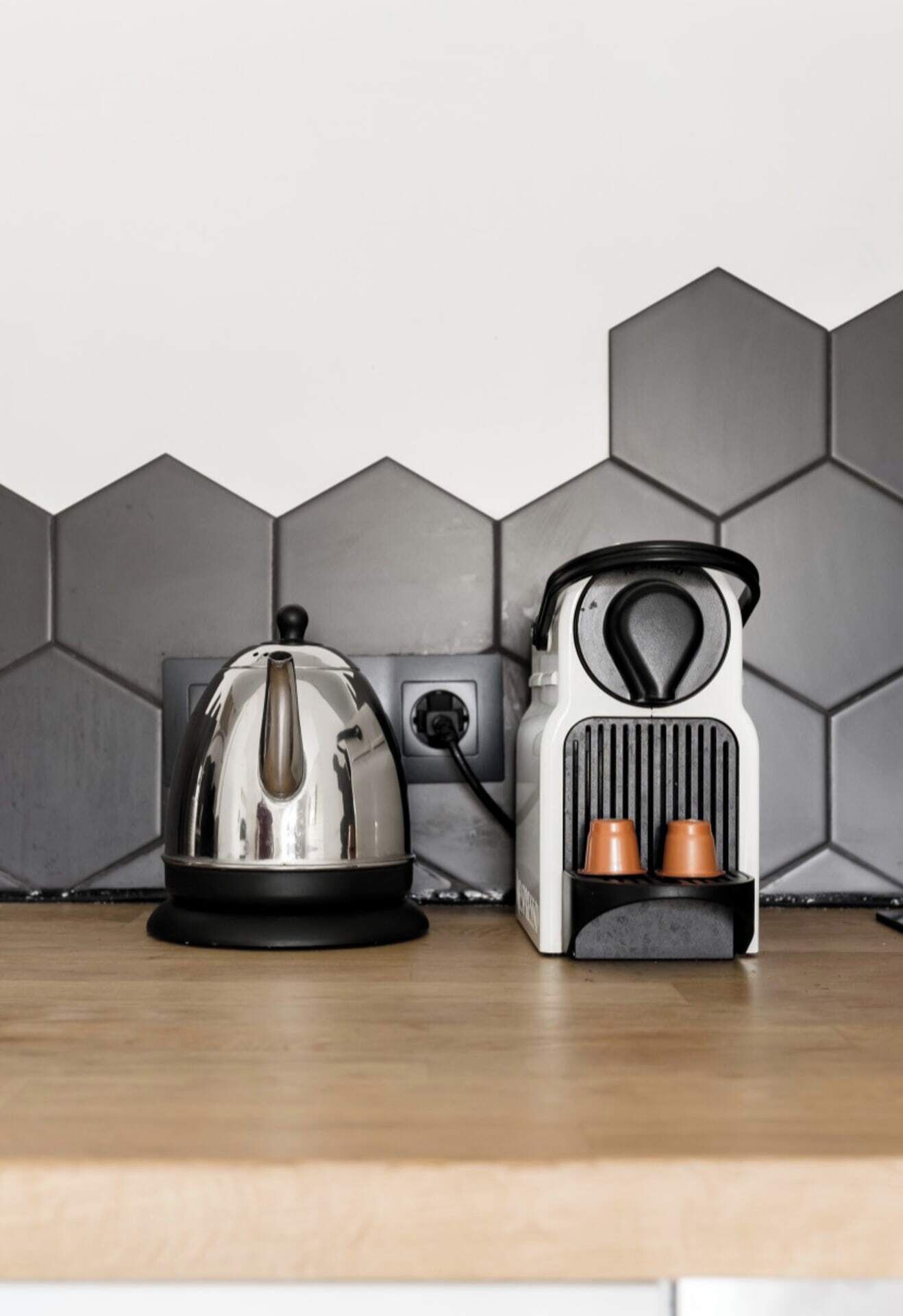Una cocina con hervidor y tostadora sobre una encimera de madera, frente a una pared con baldosas hexagonales.