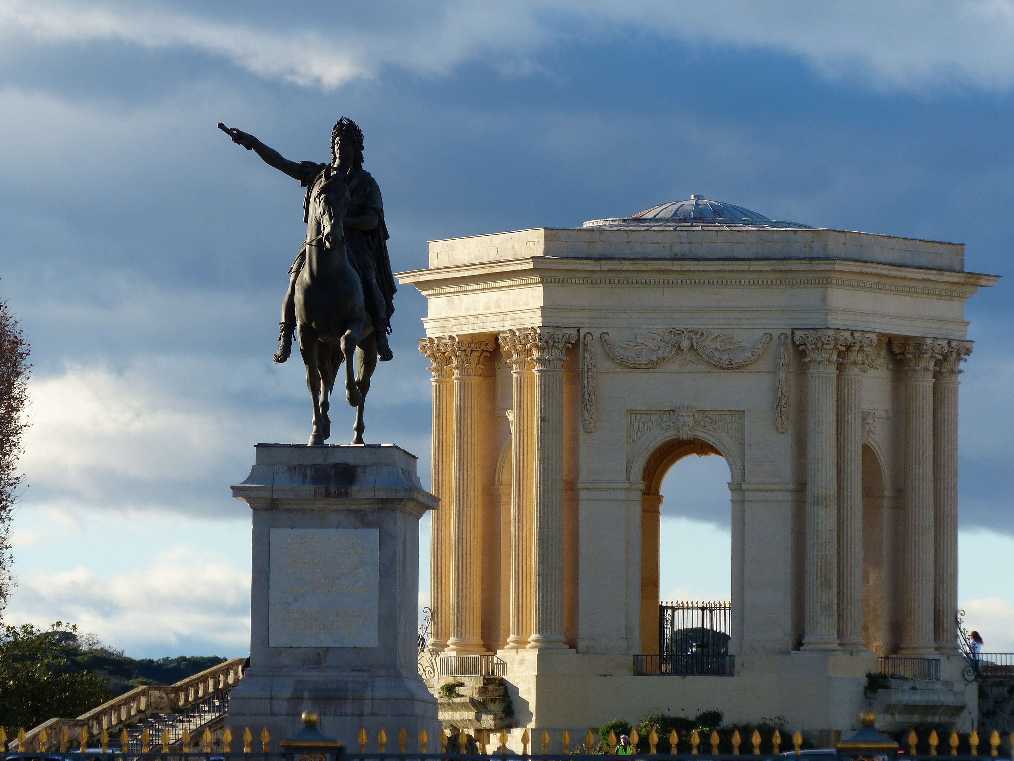Une statue représentant un homme à cheval, placée à côté d’un bâtiment de style classique, doté de colonnes et d’une arche.