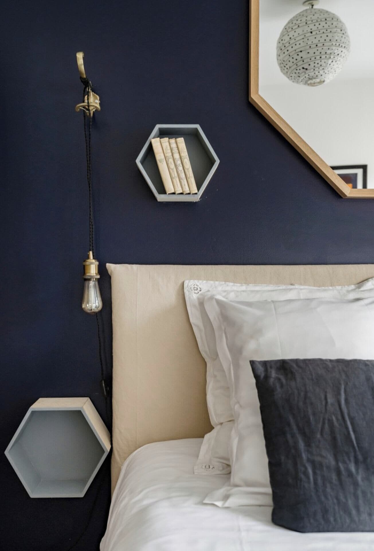 Un dormitorio con una pared azul oscuro, un estante hexagonal con libros, una lámpara colgante y un cuadro enmarcado.