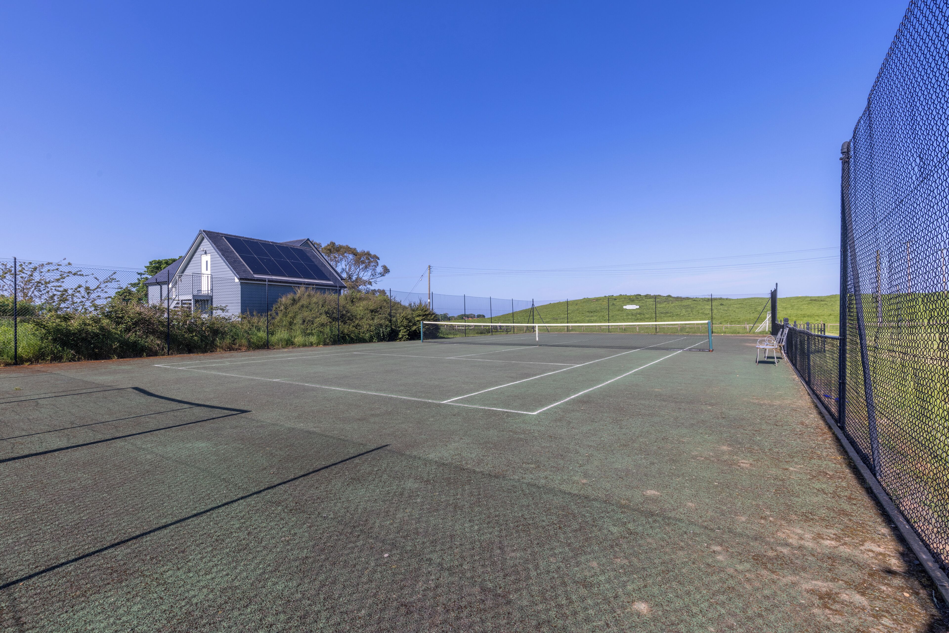 An outdoor tennis court with a net, surrounded by a fence and a house with solar panels.
