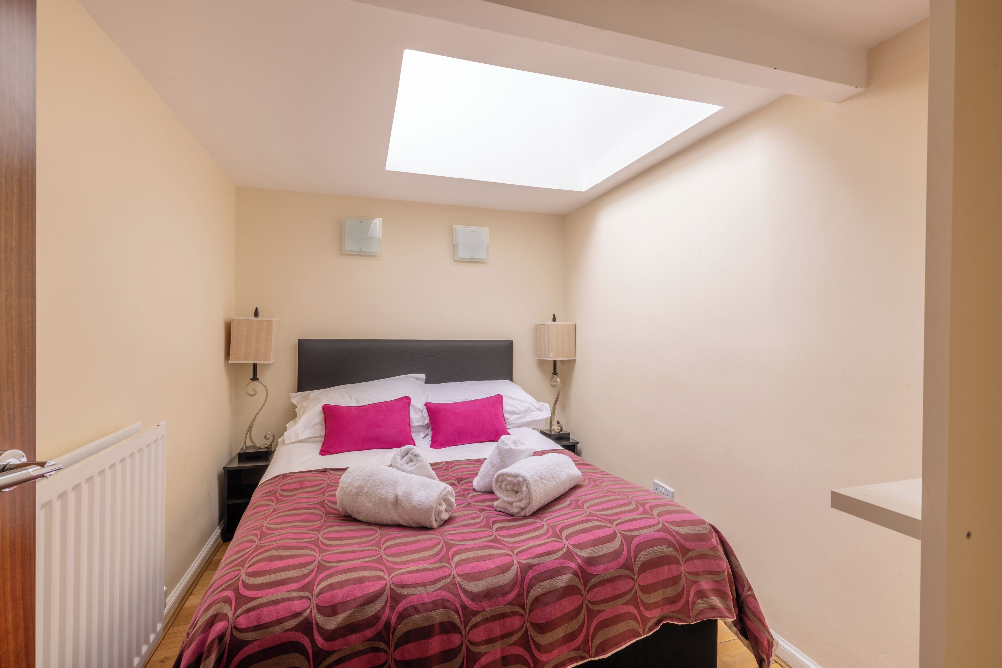 A bedroom with a skylight, a bed with pink pillows, two bedside lamps, and two rolled towels on the bed.