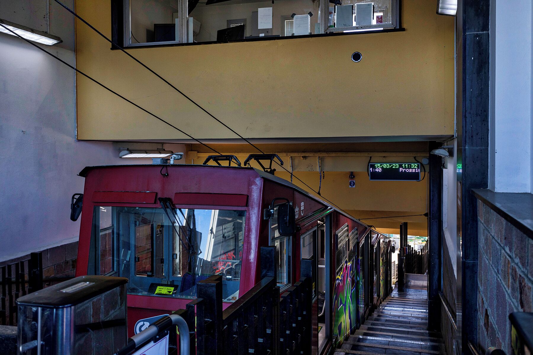 A train station with a red train, a digital display showing the time and destination, and a staircase leading to an upper level.