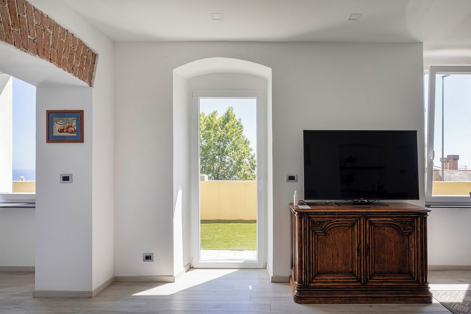 A spacious living room with a large TV, a wooden cabinet, and a view of a green lawn through an open archway.