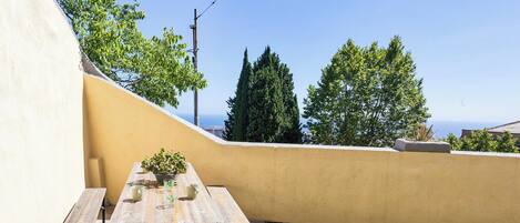 Un patio con mesa y sillas de madera, una planta pequeña en maceta y vistas al mar y a los árboles.