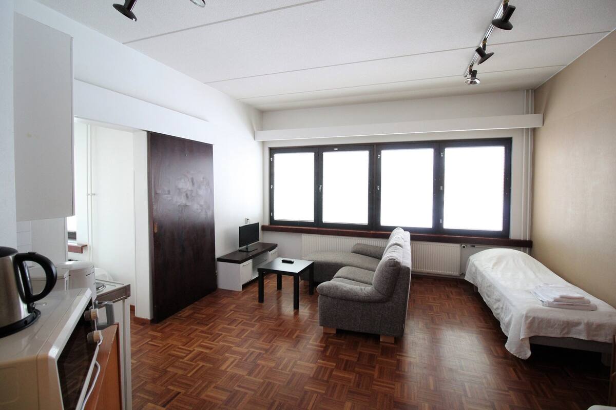 Habitación compacta con kitchenette, sofá, cama individual y un amplio ventanal que proporciona luz natural.