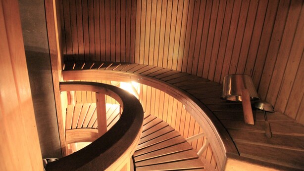 A spiral wooden staircase in a sauna room with a wooden bench and a metal container on the floor. - Forenom Apartments Rauhankatu 16 (Lahti)
