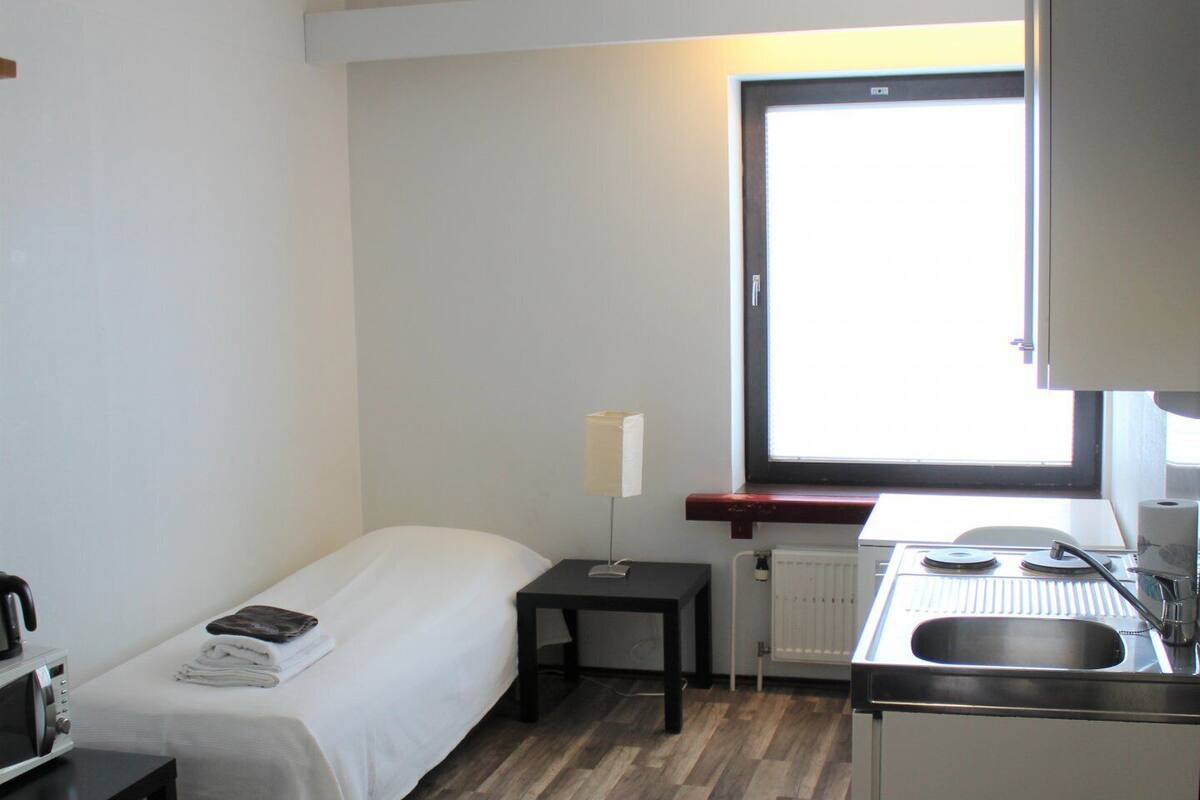 Habitación de hotel compacta con una cama individual, un escritorio pequeño, kitchenette y ventana que proporciona luz natural.