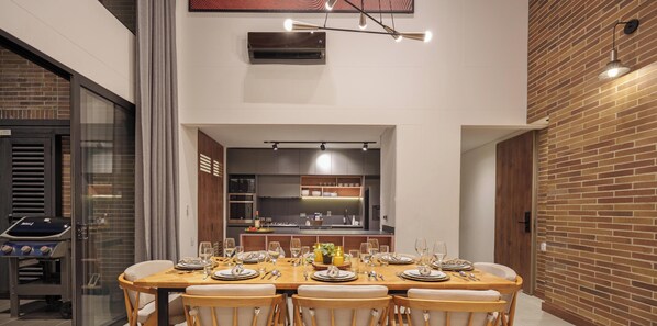 A dining area with a long table set for six, a kitchen in the background, and a brick wall on one side.
