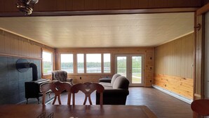 A cozy cabin interior with a fireplace, wooden walls, a dining table with chairs, and large windows offering a view of a lake.