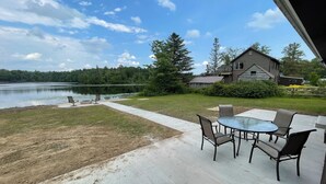 A patio with outdoor furniture overlooking a lake, a wooden house, and a grassy area.