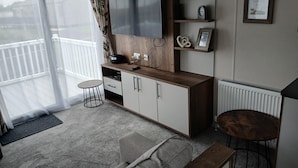 A modern living room with a large flat-screen TV, wooden cabinets, a small round table, a grey sofa, and a balcony with a white railing. - 3-bed Caravan - Haven Lakeland Leisure Park (Grange-over-Sands)