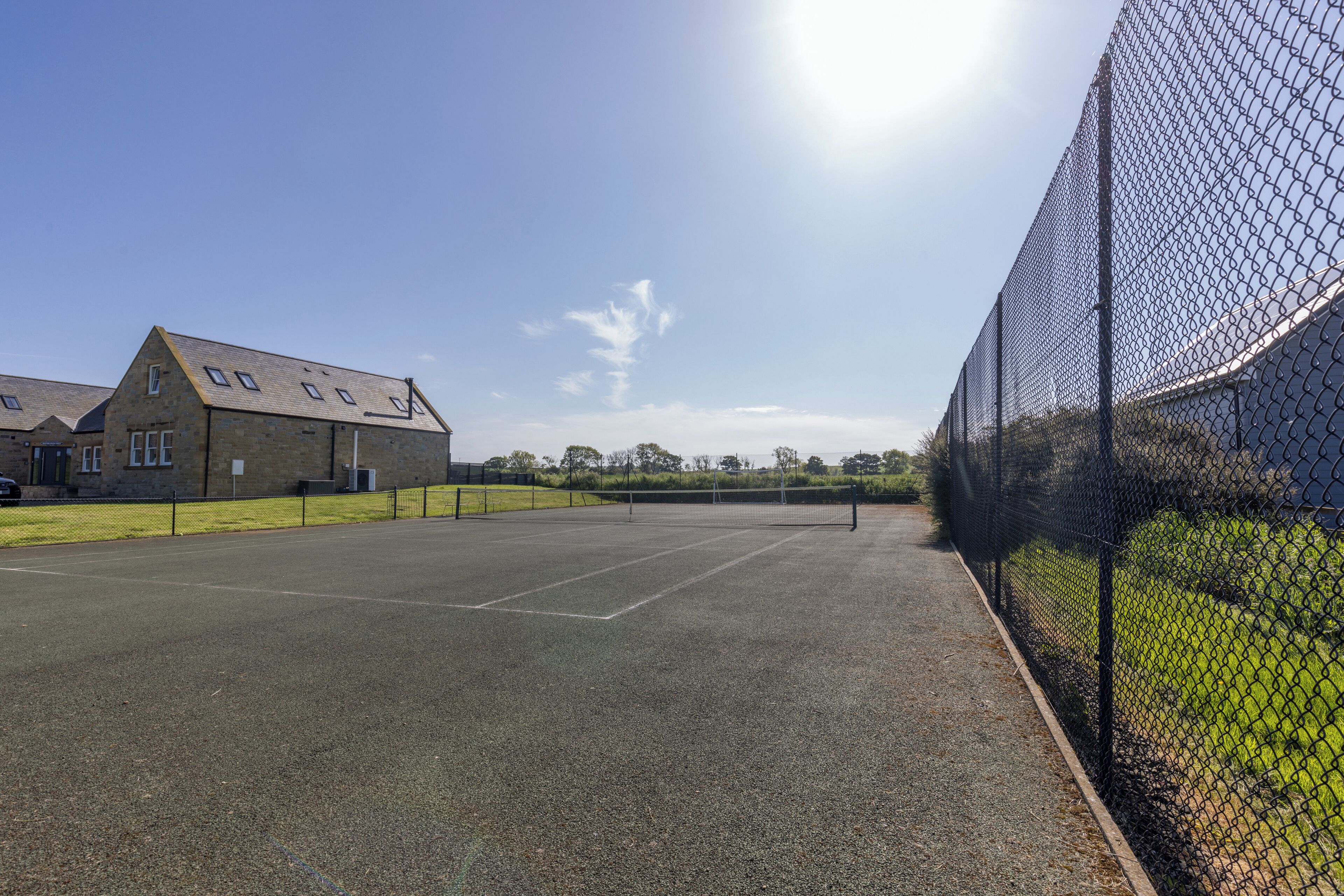 Un court de tennis entouré d’une clôture en treillis métallique, un bâtiment doté d’un toit en pente et un ciel dégagé.