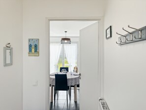 A dining area with a table, chairs, and a view through a window with curtains. - Le Réséda, Maison Avec Jardin (Saint-Valery-sur-Somme)