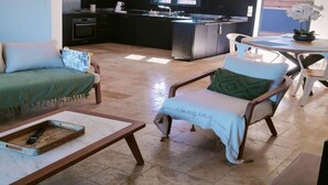 Ein Wohnzimmer mit einer Couch, einem Lounge-Sessel, einem Couchtisch und einer Küche im Hintergrund.