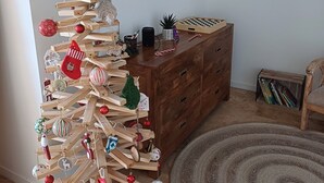 Ein Holz-Weihnachtsbaum mit Dekorationen und einer kleinen Figur, eine Holzkommode, ein Bücherregal und ein runder Teppich.