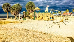 A beach playground with swings and a play structure, palm trees, and a beach in the background.