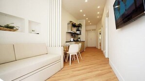 A modern living room with a white sofa, a dining area with a small table and chairs, and a kitchen in the background.