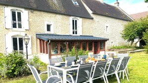 Una casa in pietra con un'area pranzo esterna coperta, un giardino e un'ampia finestra con persiane.