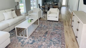 A spacious living room with a white sofa, two armchairs, a coffee table, and a patterned rug. A kitchen with white cabinets and a dining area is visible in the background.