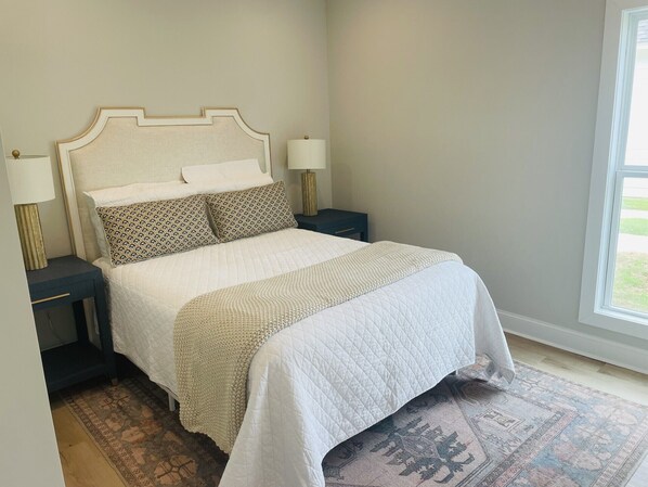 A neatly made bed with a quilted white cover, two patterned pillows, and a headboard with a decorative frame. There are two bedside tables with lamps, a patterned rug on the floor, and a window with a view of the outdoors.