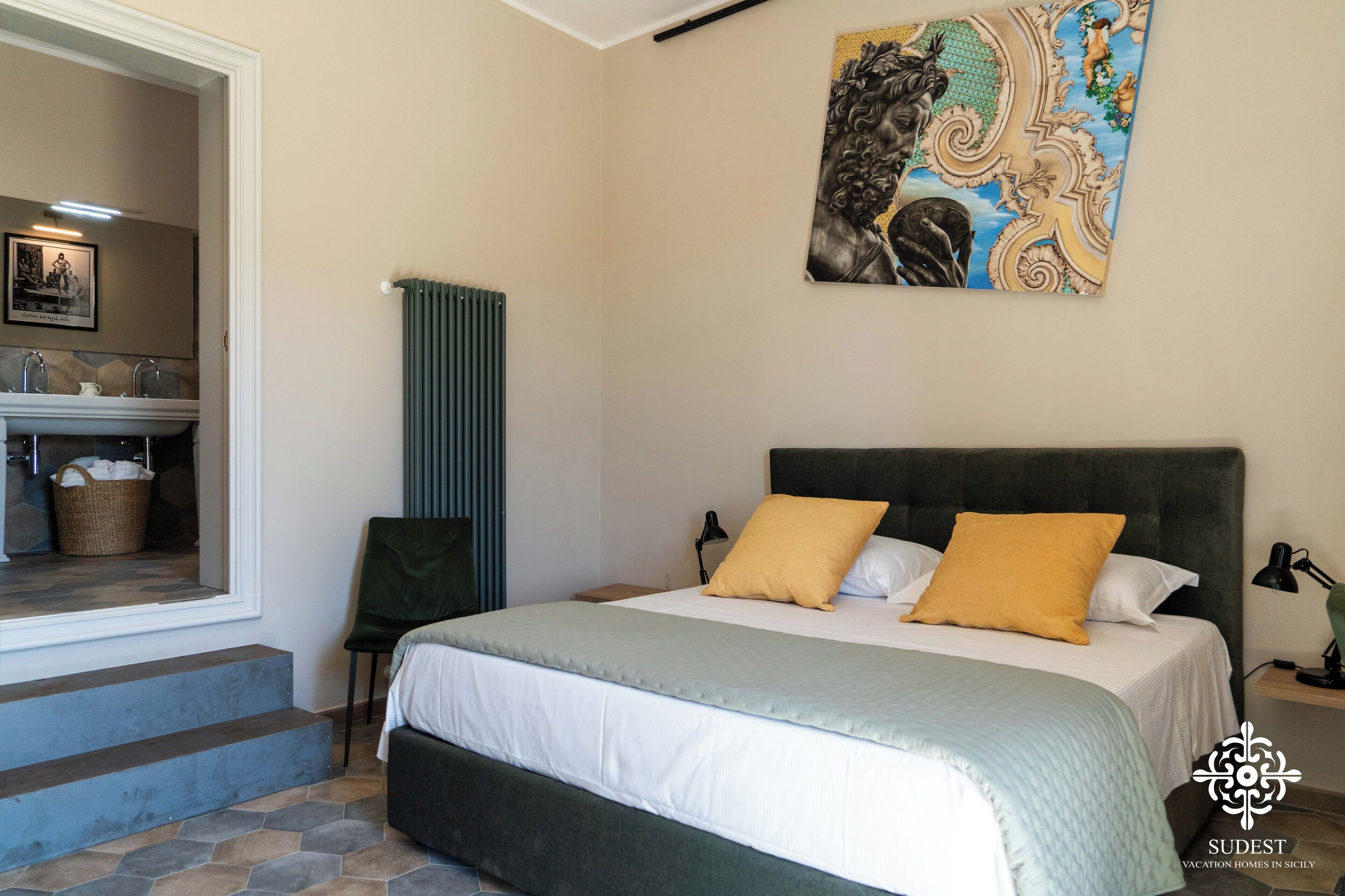 A bedroom with a bed, a chair, a radiator, a painting, and a view of a bathroom.