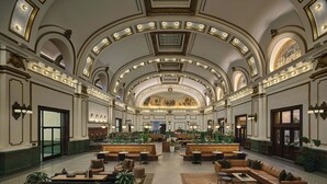Un vestíbulo amplio con techo alto, detalles ornamentales en el techo y una zona de estar central con sofás y mesas.