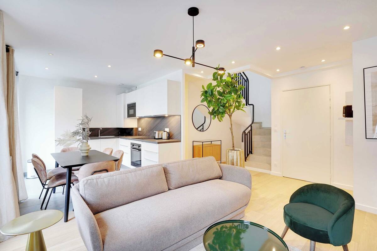 Una sala de estar moderna con un sofá, una mesa de comedor y un sillón verde. Se ve una escalera que sube a otro nivel.