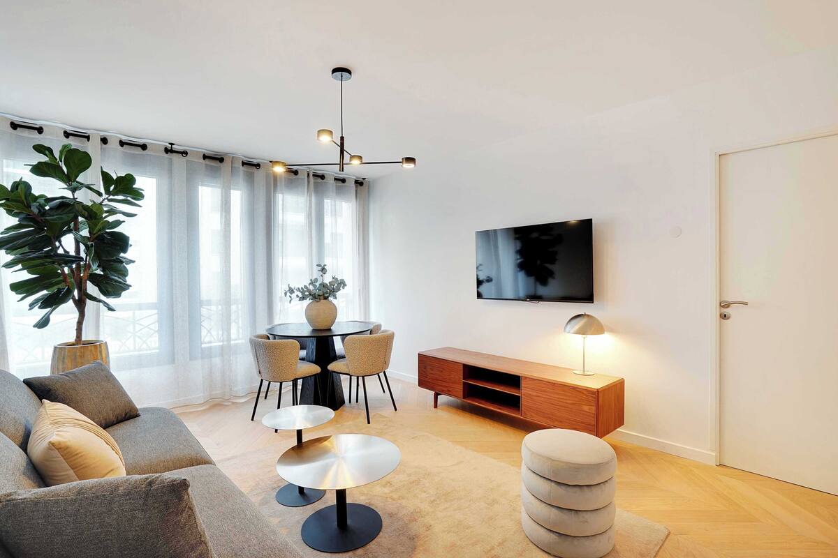 Una sala moderna con sofá, mesa redonda de centro, mesa de comedor con sillas, una planta en maceta, televisor montado en la pared y un mueble de madera.
