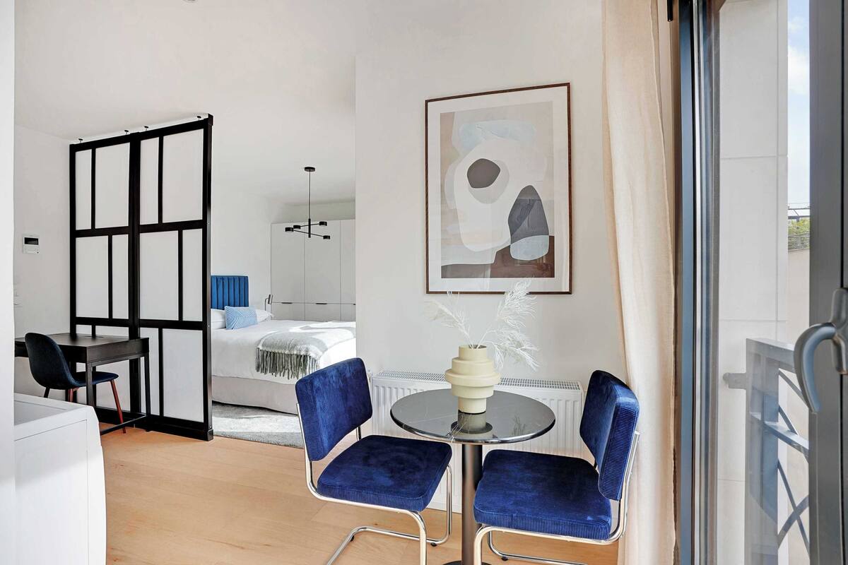 Una habitación de hotel moderna con cama, escritorio con silla, una mesa redonda con dos sillas, radiador y un cuadro abstracto enmarcado en la pared.