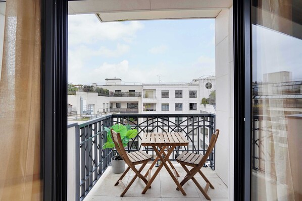 A balcony with a wooden table and chairs, a potted plant, and a view of buildings in the background.
