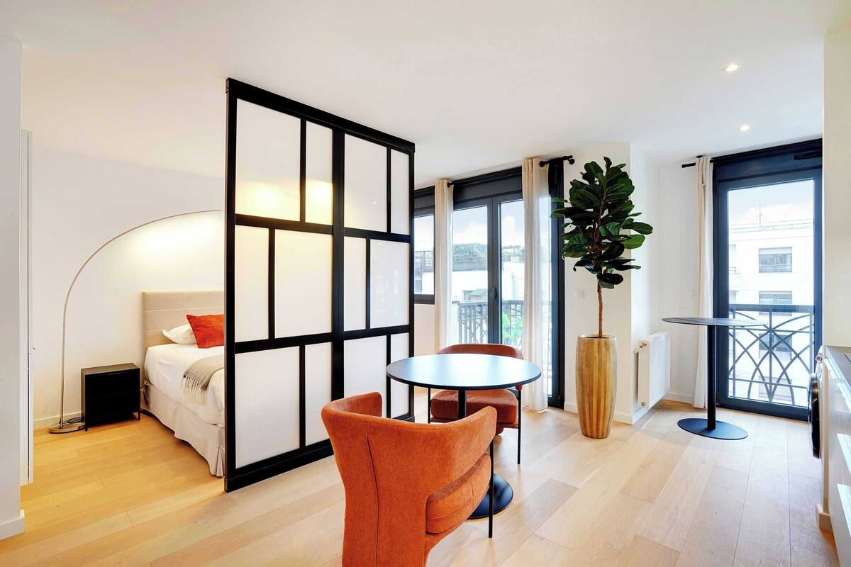 Una habitación de hotel moderna con una cama, una mesa redonda, dos sillas, una planta en maceta y ventanas amplias.