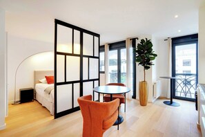 A modern hotel room with a bed, a round table, two chairs, a potted plant, and large windows.
