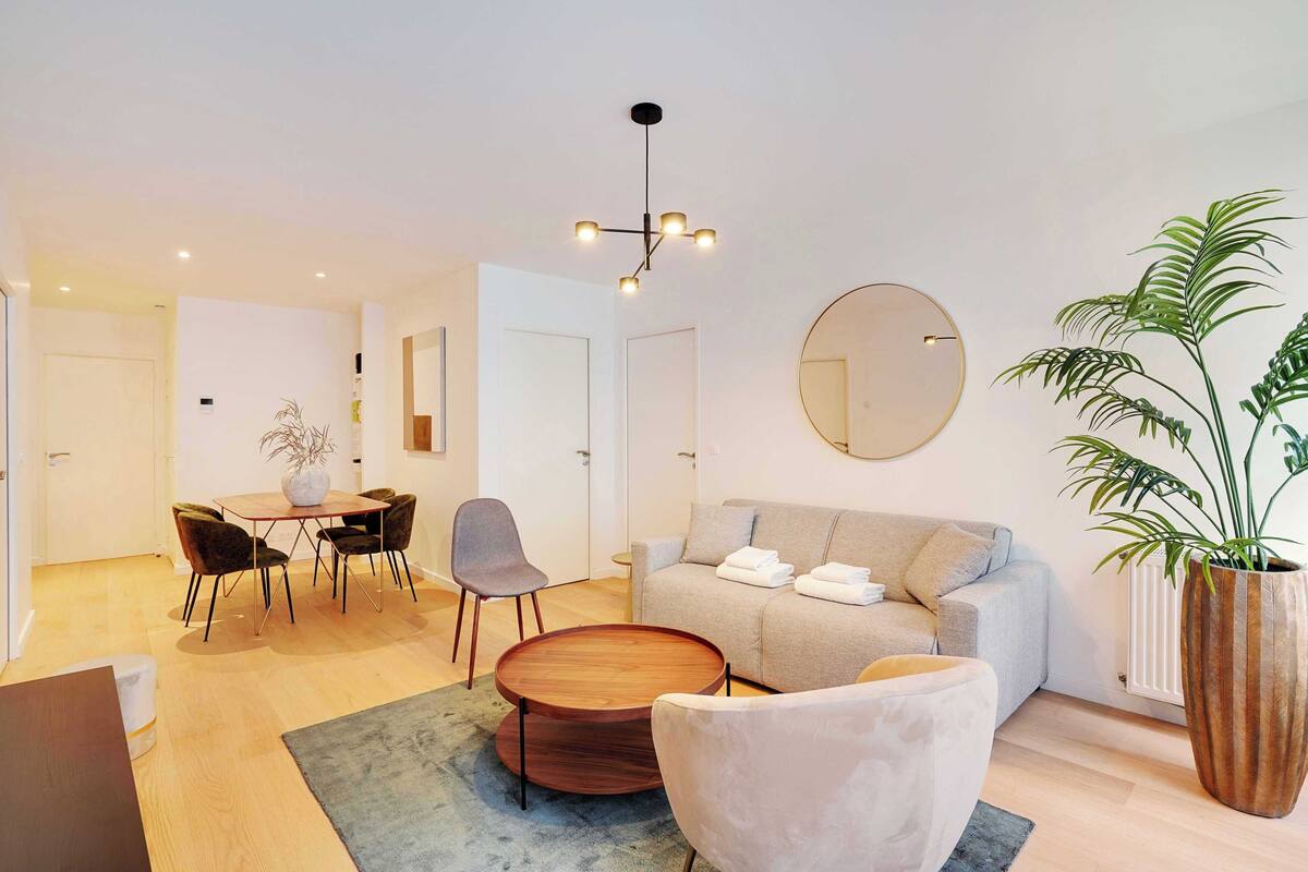 Una sala de estar moderna con un sofá, una mesa de centro y sillas. Hay una zona de comedor con una mesa y sillas. Una planta grande está en la esquina.